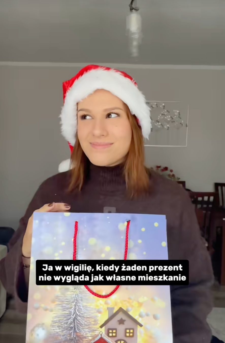Kadr z filmu Dynamic Development — kobieta w czapce Mikołaja z prezentową torbą, napis „Ja w wigilię, kiedy żaden prezent nie wygląda jak własne mieszkanie”
